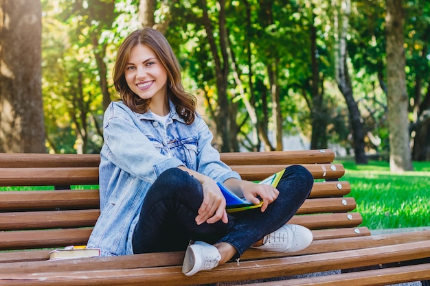 Young girl student sits on a bench in the park and holds books, notebooks.