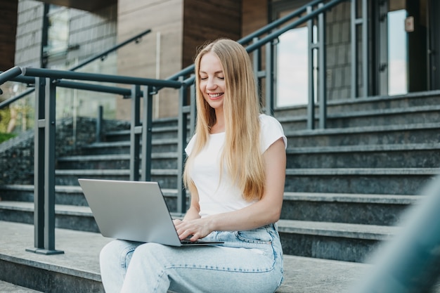 Young girl student sitting with laptop on stairs near university smiling and studying