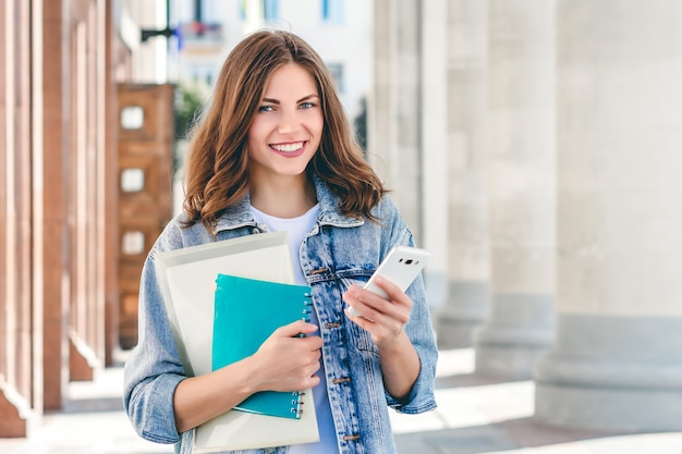 若い女子学生が大学に対して笑顔します。かわいい女子学生は、フォルダー、ノートブック、携帯電話を手に保持しています。学習、教育