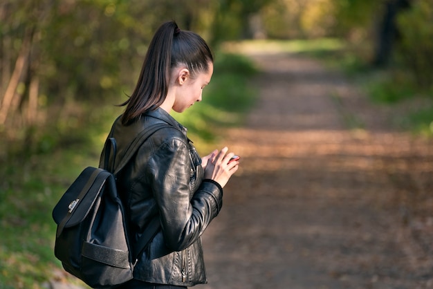 若い女の子の学生は彼女の手で電話を持って公園に立っています。ポニーテールの女の子が公園を歩いています。背面図。