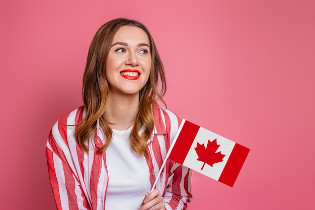 Young girl student wearing red striped shirt smiling and holding a small canada flag and looking away isolated over pink space, Canada day celebration