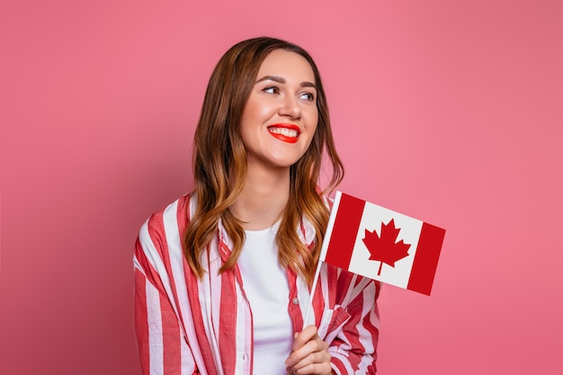 Young girl student wearing striped shirt smiling and holding a small canada flag isolated over pink space, Canada day