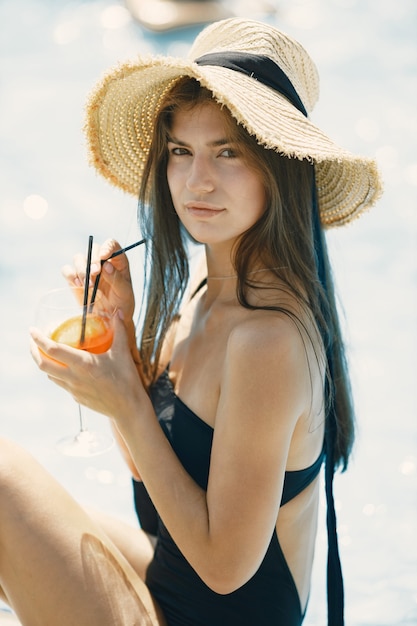 Young girl in a summer hat sitting near swimming pool. Girl wearing black swimwear and a hat