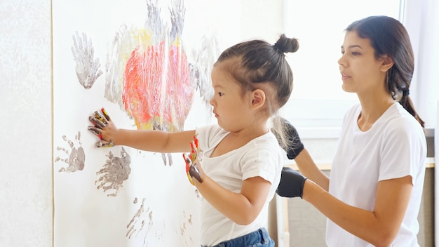 Young girl in t-shirt and jeans plays with mother drawing on large white canvas with hands standing in studio against bright window