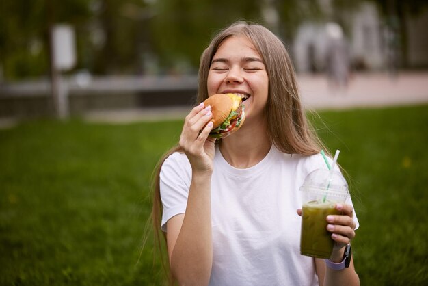 A young girl takes out a burger from a paper bag sitting on the green grass the