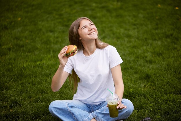 若い女の子が緑の草の上に座って紙袋からハンバーガーを取り出す