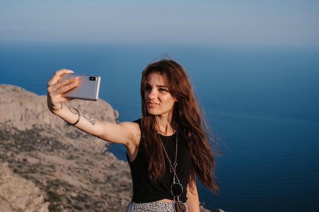 A young girl takes a selfie on her smartphone against the background of the sea at the cliff The girl smiles at the camera The concept of tourism recreation