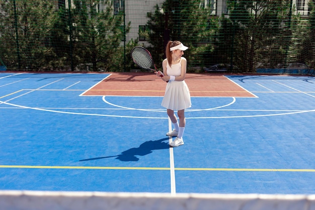 白いユニフォームの若い女子テニス選手がテニスコートでラケットを握り女性アスリートがプレーしています