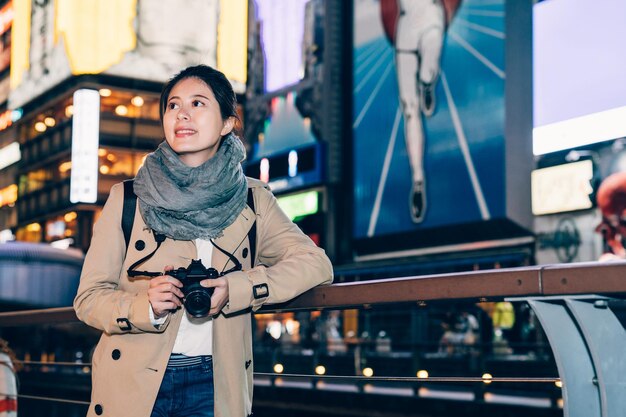 young girl travel photographer cheerfully smiling looking up to the building standing nearby the canal in dotonbori osaka japan. lady toursit holding camera relying on handrail at dark night city.