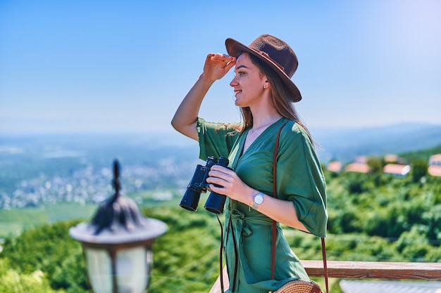 Young girl traveler wearing hat holds binoculars and looks into the distance during vacation weekend trip on a bright sunny day