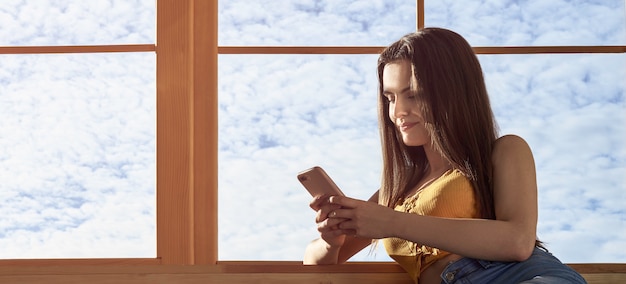 young girl using smartphone and setting on window sill with copy space