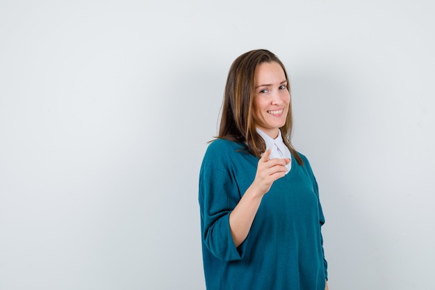 Young girl in v-neck sweater, shirt pointing at front, standing sideways and looking glad.