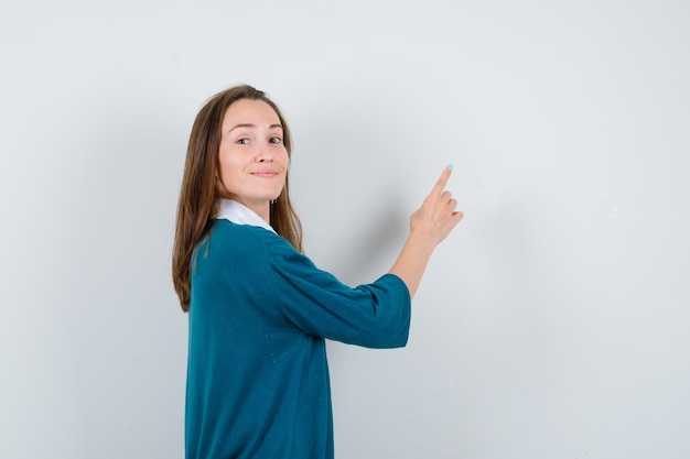 Young girl in v-neck sweater, shirt pointing up, standing sideways and looking pleased .