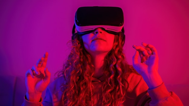 Young girl in virtual reality glasses with blue and red illumination in the room with rised up hands. Entertainment at home