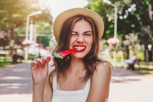 A young girl walks in the park with a lollipop laughs and grimaces. Charming girl eats lollipop and wrinkles her nose