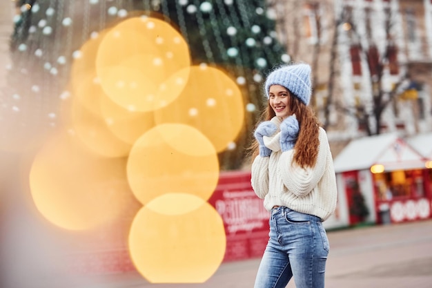 Молодая девушка в теплой одежде гуляет по городу на фоне рождественской елки.