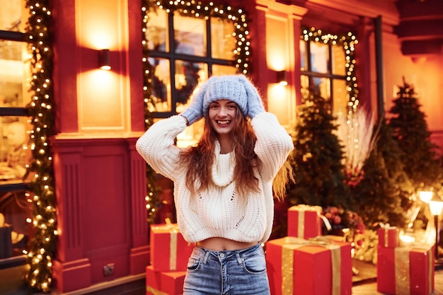 Young girl in warm clothes have a walk outdoors in the city at evening time near building with artificial festive lights.