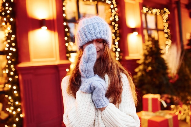 Young girl in warm clothes have a walk outdoors in the city at evening time near building with artificial festive lights.