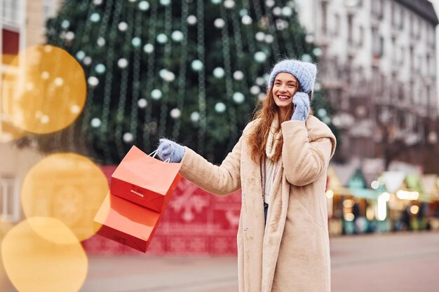 Photo young girl in warm clothes with shopping bags in hand have a walk outdoors in the city and holding phone.