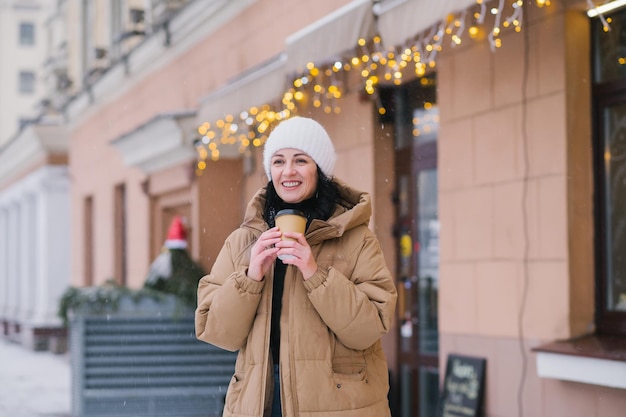 A young girl in a warm hat and coat drinks tea outside in winter near a cafe and laughs