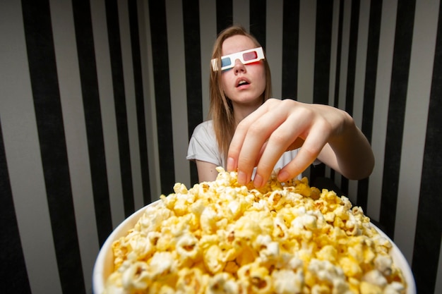 Молодая девушка смотрит фильм и ест попкорн в 3D-очках на фоне полосатой стены
