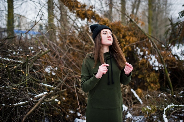 Young girl wear on long green sweatshirt jeans and black headwear at branches of the pine tree in winter day