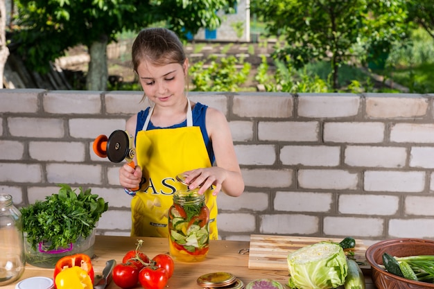 エプロンを身に着けている少女は、保存または酸洗いのために新鮮な野菜を準備し、庭の近くに立って蓋を締めるツールを持っています