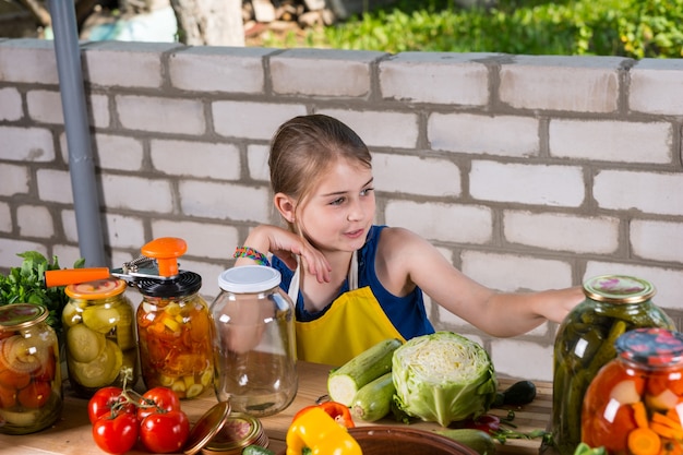 庭のレンガの壁の近くに立ってエプロンを着て、ガラスの瓶で保存または酸洗いのために新鮮な野菜を準備している若い女の子