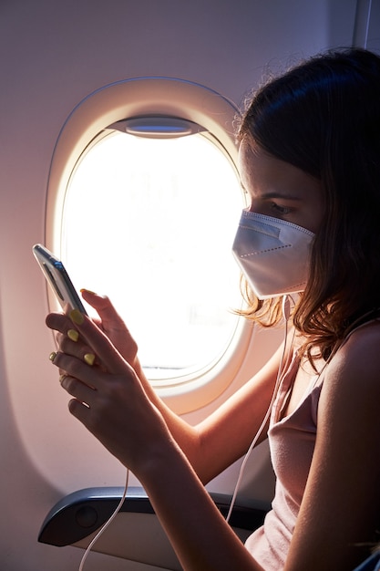 Foto giovane ragazza che indossa la maschera per il viso utilizza lo smartphone durante il viaggio in aereo