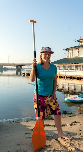 A young girl wearing a full cap, blue t-shirt and board short\
stands with a paddle