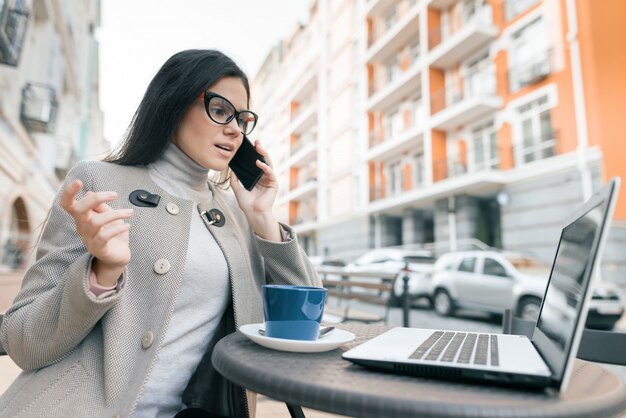 屋外カフェに座っている暖かいコートで眼鏡をかけている若い女の子