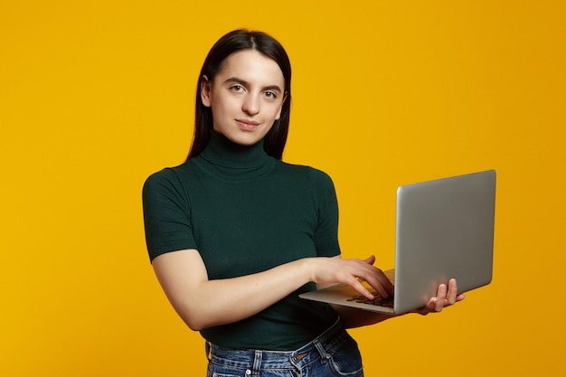 Young girl wearing green tshirt standing while using laptop device isolated on yellow background