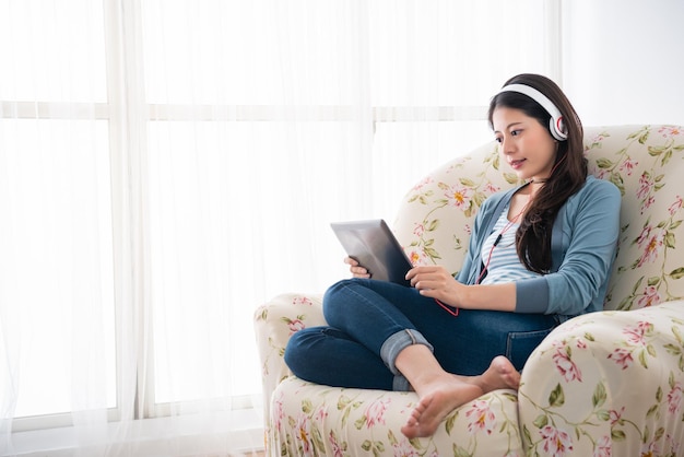 young girl wearing headphone and watching learning videos on tablet sitting on the couch joyfully.