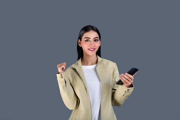 灰色の背景のインドのパキスタンのモデルに携帯電話を保持している正面を見てジャケットを着ている若い女の子