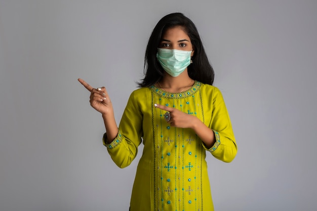 Young girl wearing a medical face mask posing, pointing and showing something on grey background.