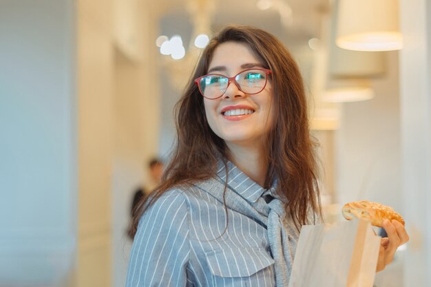 Young girl wearing red glasses holding pastry with paper bag in bakery store