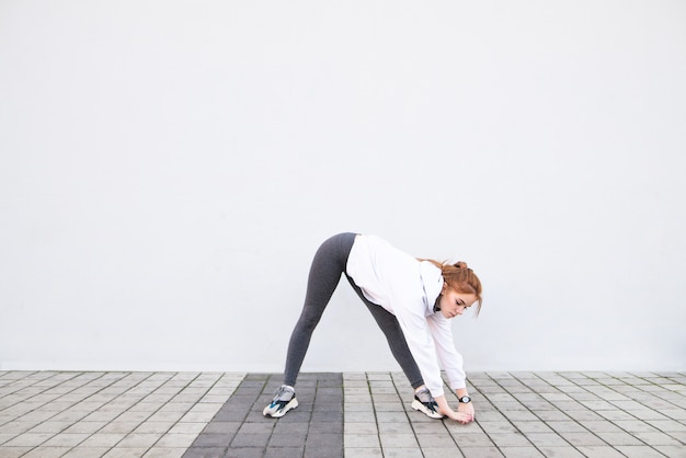 Young girl wearing sportswear stretches, makes stretchings on the background of a white wall.