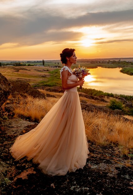 Ragazza in un abito da sposa fiutando un bouquet da sposa al tramonto