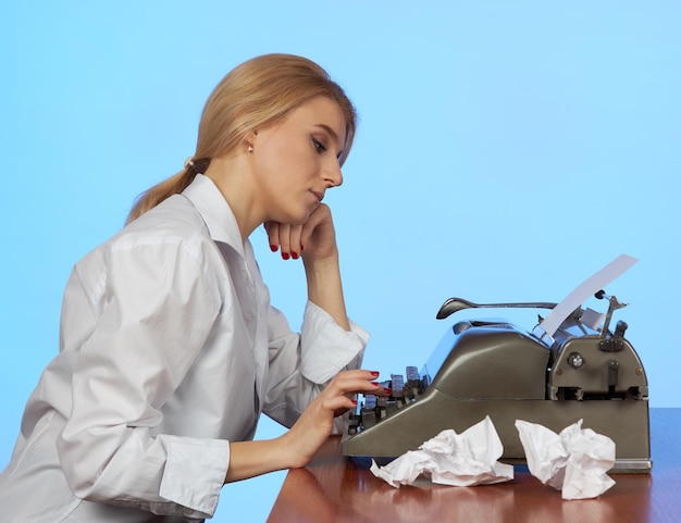 A young girl in a white blouse sits at a desk in the office and types text on a retro typewriter.