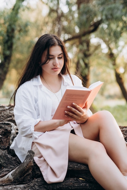 Young girl in white clothes sits on a tree in the forest and reads a book