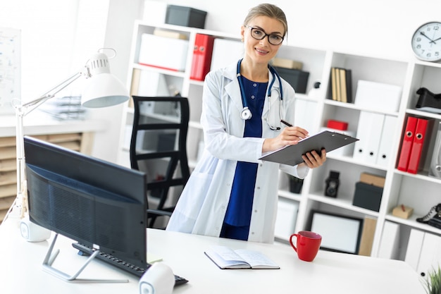 A young girl in a white coat is standing near a table in her office and holding a tablet and a pen