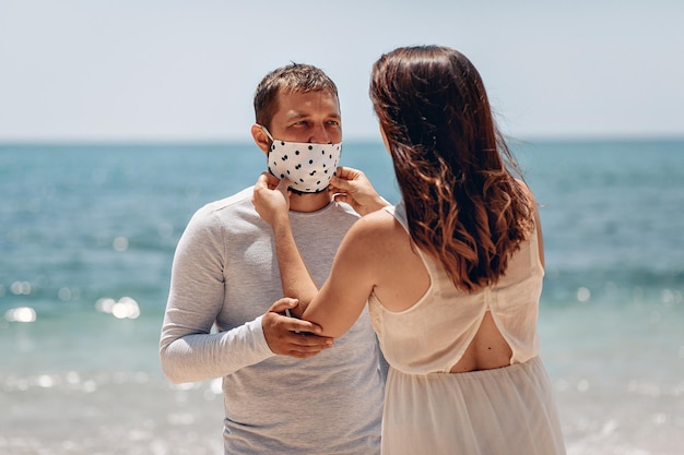 Young girl in a white dress carefully putting a protective medical mask with black polka on her husband to protect him from the virus. Family concept