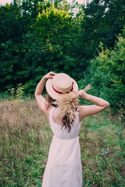 Young girl in a white dress and hat on a field background. The girl in a hat stands with her back to a bouquet of dried flowers.