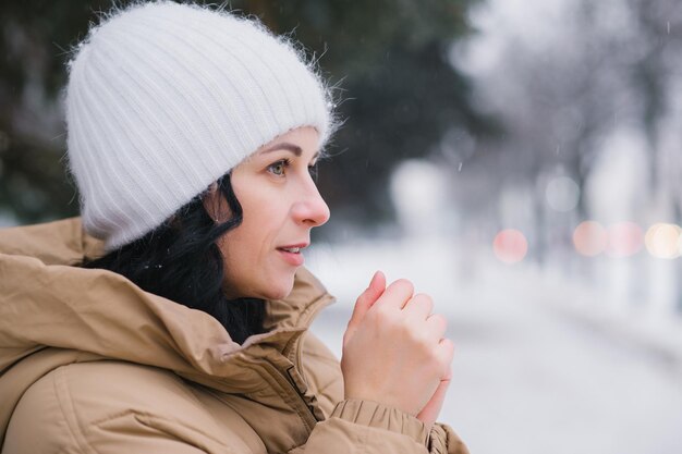 A young girl in a white hat warms her hands It stands in the city in winter under the snow