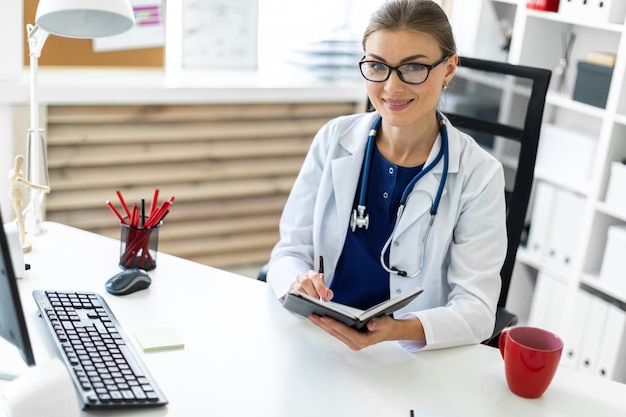 A young girl in a white robe sits at a table in the office and holds a pen and a notebook in her hand. a stethoscope hangs around her neck.