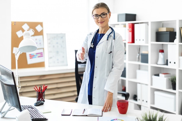 A young girl in a white robe stands near the desk in the office and stretches her right hand forward. 