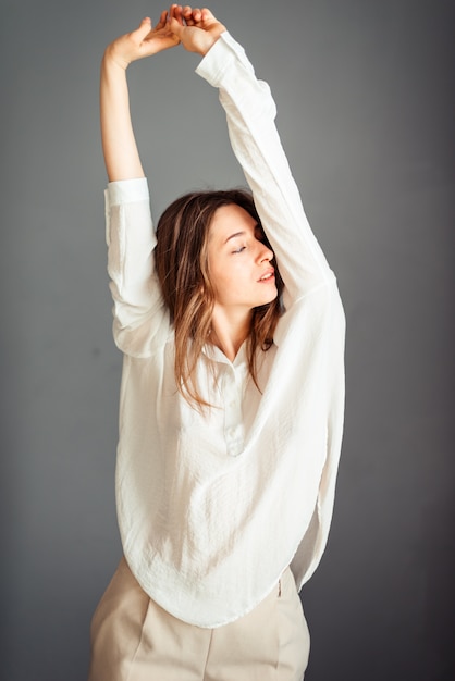 A young girl in a white shirt, both hands are straight at the top, pulls, stretches up, isolator on a gray wall. No retouching. Without makeup.