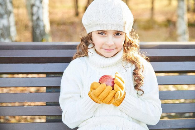 A young girl in a white sweater and beret sits on a bench in the park in autumn with an apple in her hands. High quality photo