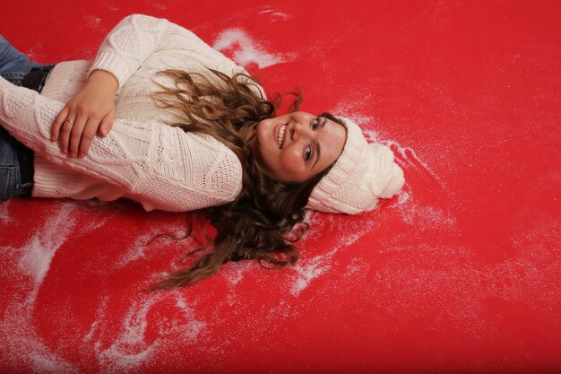 Photo a young girl in a winter hat and a knitted sweater lies on a red background artificial snow