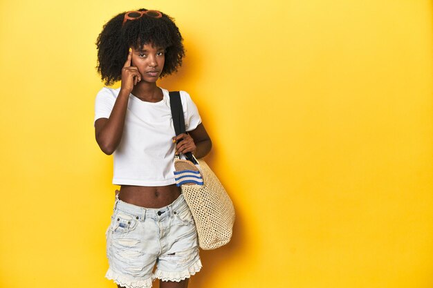 Young girl with beach bag ready for holiday yellow backdrop pointing temple with finger